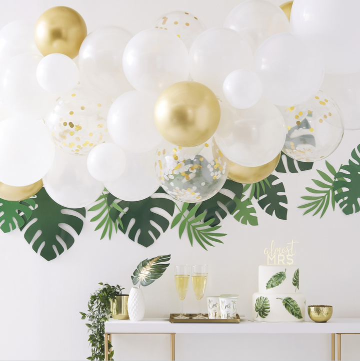 Tropical-themed bridal shower setup with matte white, pearl white, gold, and clear gold confetti-filled balloon arch with green foliage accents. Faux monstera leaves and circular wooden bride to be signs on a white wall above a gold and white beverage table.