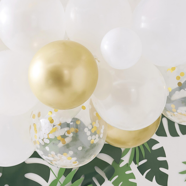 matte white, pearl white, gold, and clear gold confetti-filled balloon arch with green foliage accents against a monstera leaves patterned white wall.
