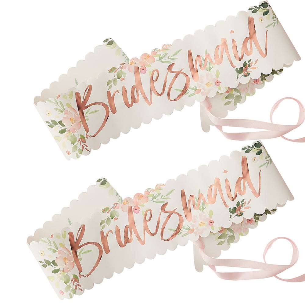 Two paper sashes with floral patterns and 'Bridesmaid' printed in rose gold foil, tied with pink ribbons.