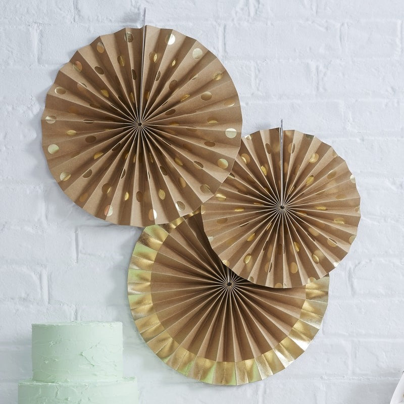 3 Kraft paper party fans with gold foil polka dot print and edge detail on a white brick wall with a mint green frosted 2-tier cake in the right lower corner.