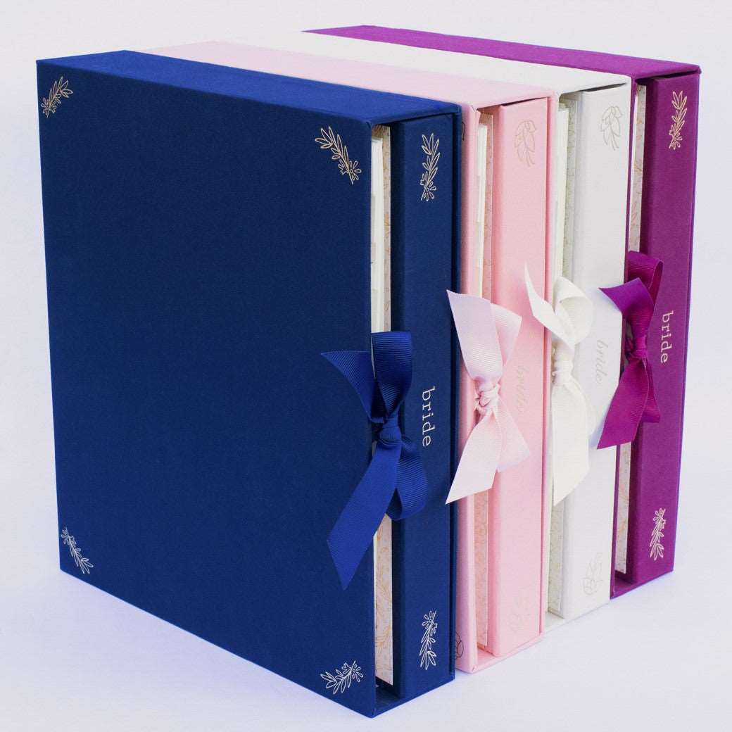 A set of fabric-covered wedding planner books in coral pink, navy, beige, and wine-burgundy on a white background