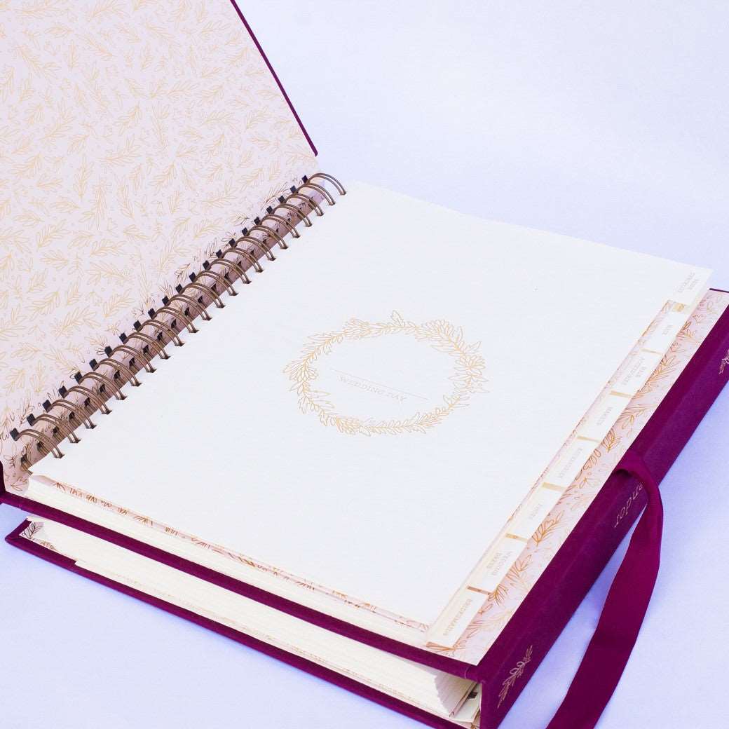 A wine-burgundy fabric-covered wedding planner book open to the introduction cover page on a white background