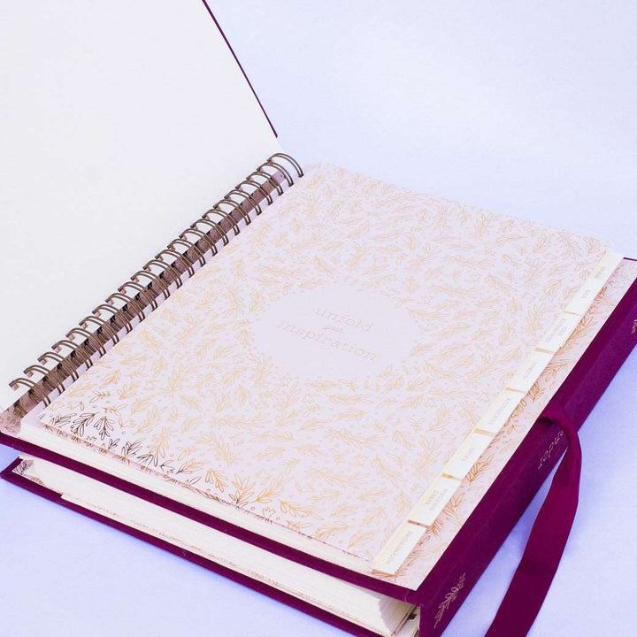 A wine-burgundy fabric-covered wedding planner book open to the inspiration section cover page on a white background