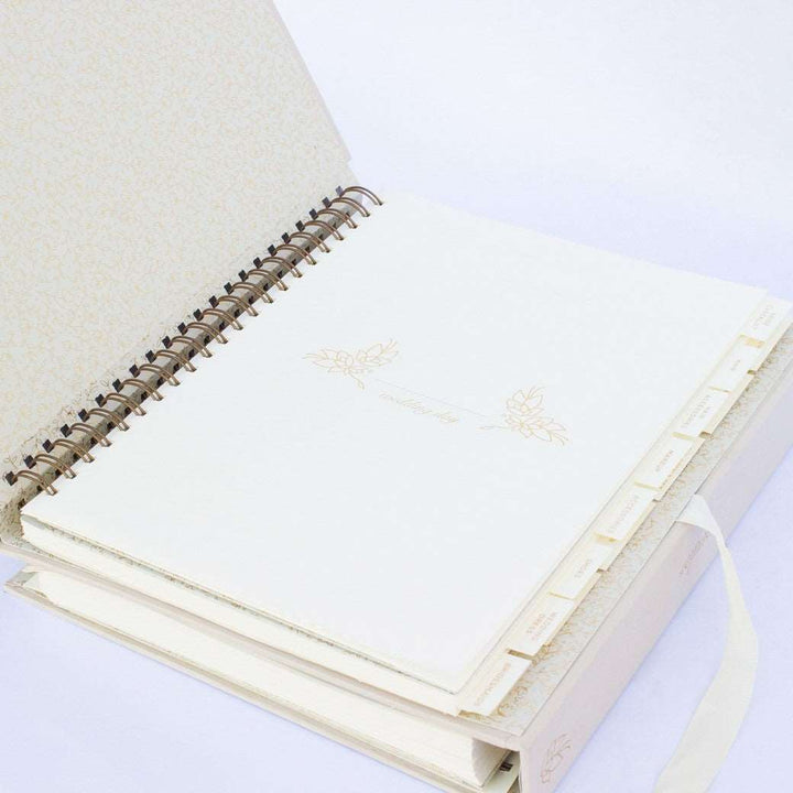 A beige fabric-covered wedding planner book open to the wedding day section cover page on a white background