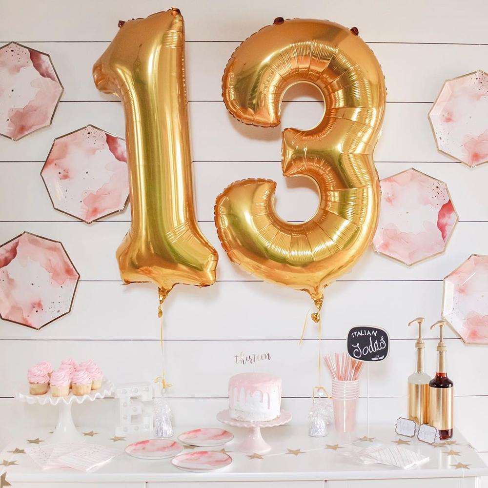 A white dessert bar with white and pink cake stands topped with pink frosted cupcakes and cakes, pink iridescent cups and matching straws, a gold star banner and syrup bottles. tonal pink, white, and gold plates decorating the wall above with gold ballons that read the number 13