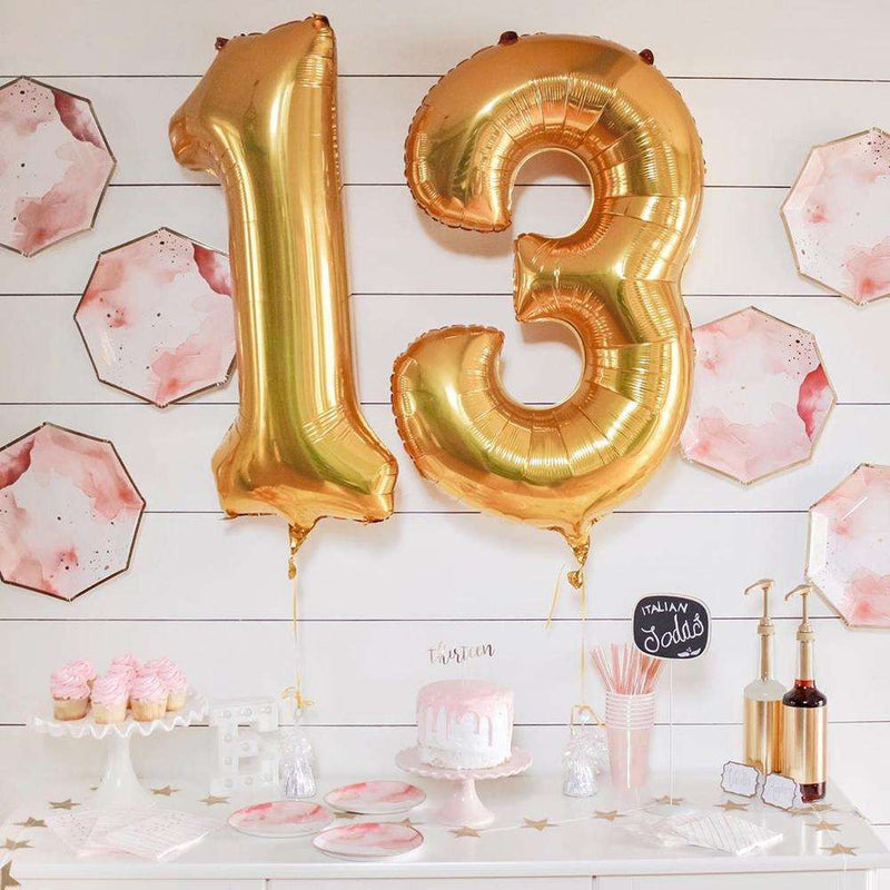 A white dessert bar with white and pink cake stands topped with pink frosted cupcakes and cakes, pink iridescent cups and matching straws, a gold star banner and syrup bottles. tonal pink, white, and gold plates decorating the wall above with gold ballons that read the number 13
