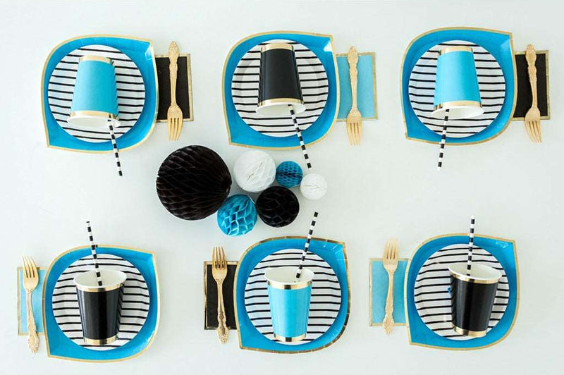 table setting of blue dinner plates with gold trim, topped with white with black stripe dessert plates, alternating settings have either a black cup with gold trim or blue cups with gold trim with black and white striped drinking straws in them and either a black napkins with gold trim or a blue napkin with gold trim topped with a gold fork