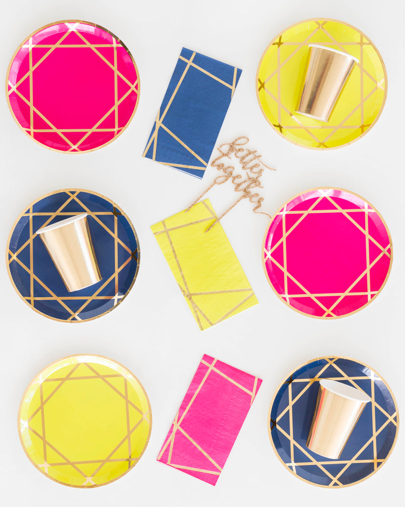A set of alternating hot pink, navy, and lemon-yellow dinner plates and napkins with gold jewel pattern print, gold paper cups, on a white background