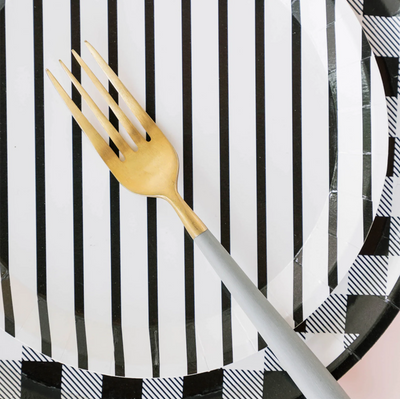 Grey handled gold fork on top of a white with black pinstripe paper plate on top of a black and white gingham printed paper plate