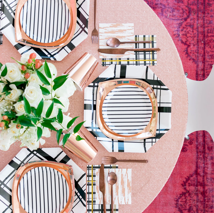 Table setting with geometric party plates in stripe, plaid, and foil finishes, cutlery, and floral arrangement on a patterned background
