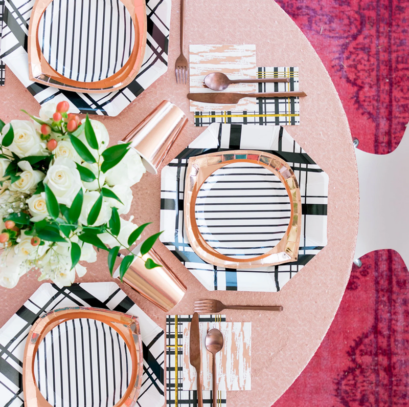 Table setting with geometric party plates in stripe, plaid, and foil finishes, cutlery, and floral arrangement on a patterned background