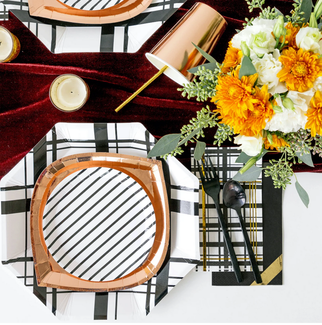 Decorative rose gold, black and white plaid, and striped table setting with floral arrangement, candles, and patterned napkins.