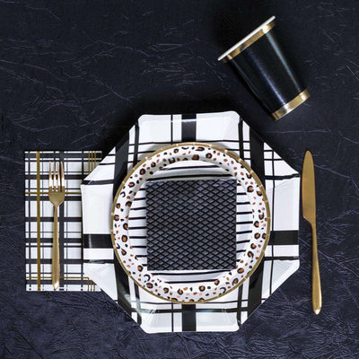 Table setting of black and white plaid printed party plate, black and white striped party plate, animal print party plate, black, white, and gold foil napkin, black and gold foil cup, black textured cocktail napkin, and gold cutlery.