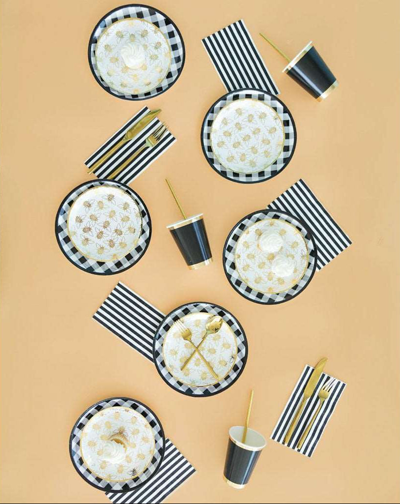 a tablesetting of black and white checker print dinner plates, topped with a white dessert plates with allover gold bumble bee print, black and white striped napkins, gold flatware sets, and black cups with gold borders on top of a and gold drinking straws.