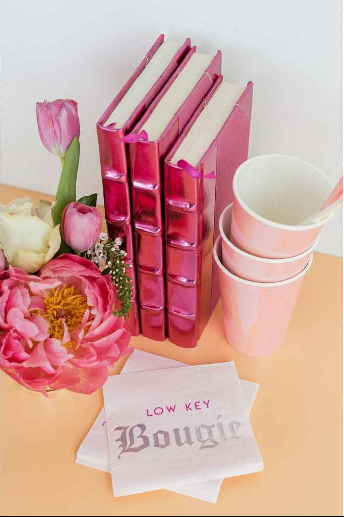 A stack of iridescent peachy pink cup with white and peach colored drinking straws next to 3 metallic hot pink covered books, a cream, lilac, green, and fucshia floral arrangements, and a stack of light lavendar napkins with the words low key bougie printed on them on a peach table