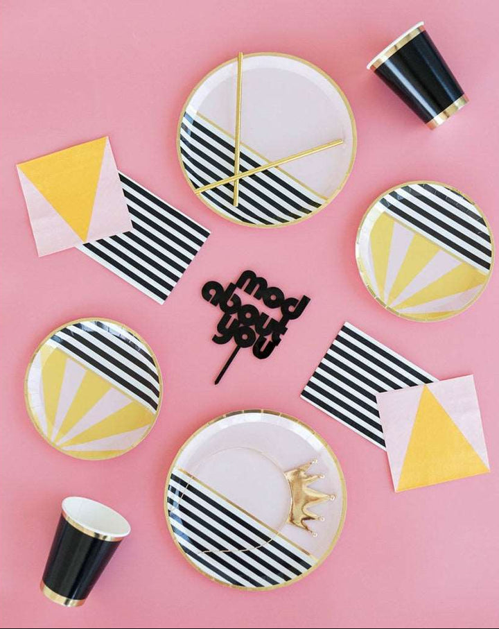 black cups with gold borders, pink napkins with yellow triangle print, white and black striped napkins, black and white striped dessert plate with pink and yellow stripe detail and gold trim, pink dinner plate with black and white striped print with gold trim, a black sign that reads mod about you, and a gold princess tiara all on a pink background.