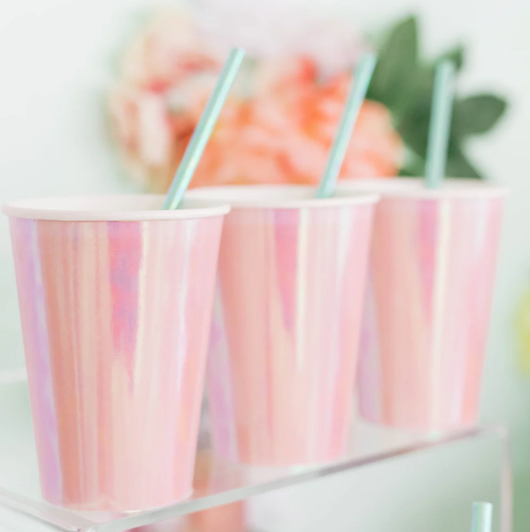 A line of iridescent peach-pink paper cups with mint green straws in them on a clear glass shelf