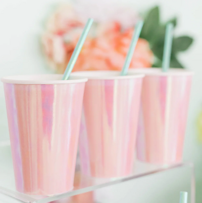 A line of iridescent peach-pink paper cups with mint green straws in them on a clear glass shelf