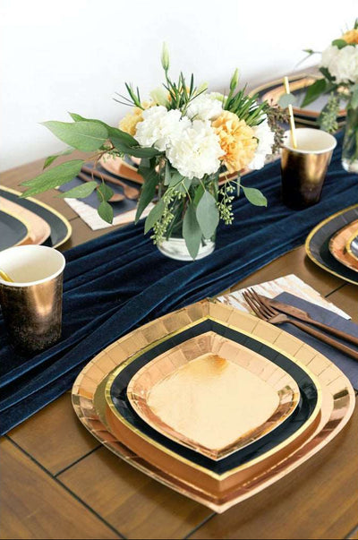 rectangle wooden table with a velvet navy runner, with yellow, white, and green floral centerpieces on the runner, table settings with gold charger plates topped with gold rimmed navy dinner plates, topped with gold dessert plates, bronze flatware, and gold paper cups
