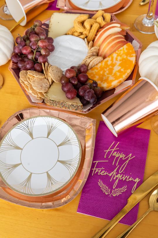 orange tablecloth with a mauve, wine, blush pink, and rose gold charger plate filled with fresh fruit, crackers, and cheese, white pumpkin decor, gold flatware, a rose gold foil dinner plate topped with a white with gold foil print dessert plate, and a purple dinner napkin with the words Happy Friendsgiving and branches printed in gold foil.