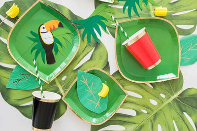 monstera printed white tablecloth topped with green dinner and dessert plates, leaf theme napkins, a toucan banner, and a black cup with gold borders and a red cup with gold borders. 