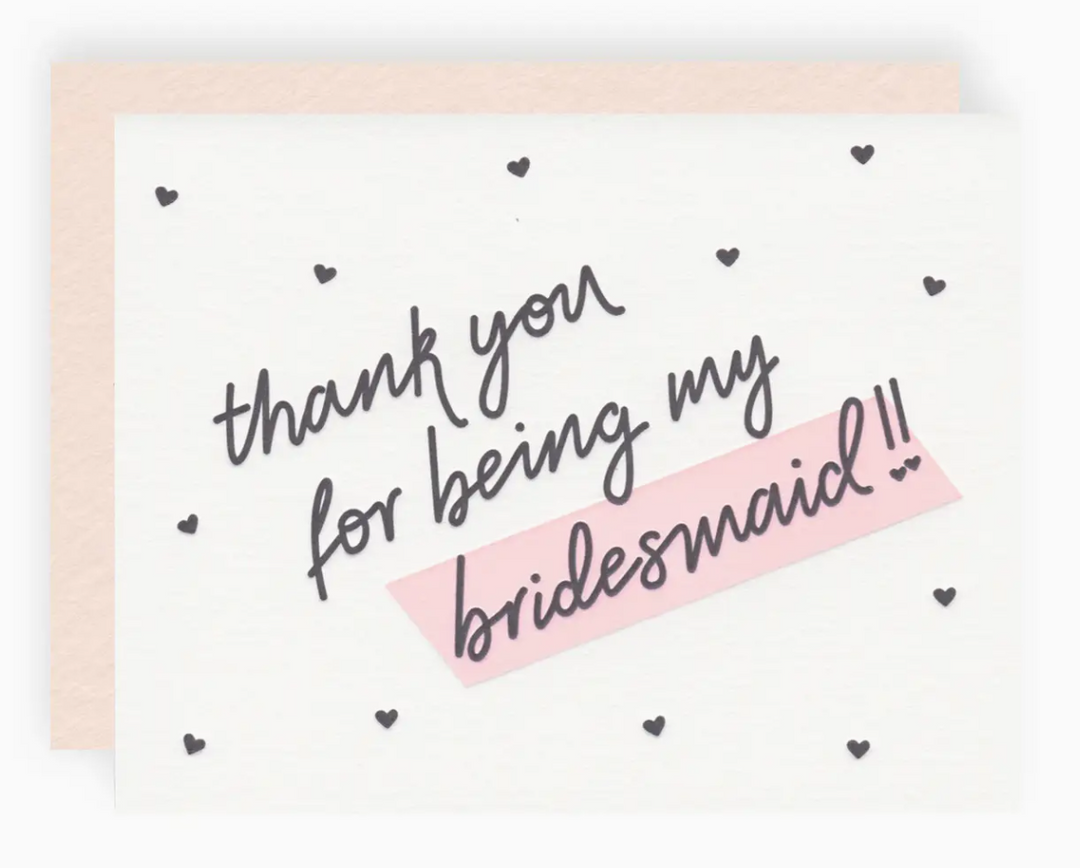 White greeting card with black mini hearts printed allover and text that reads "thank you for being my bridesmaid!!" paired with a blush pink envelope