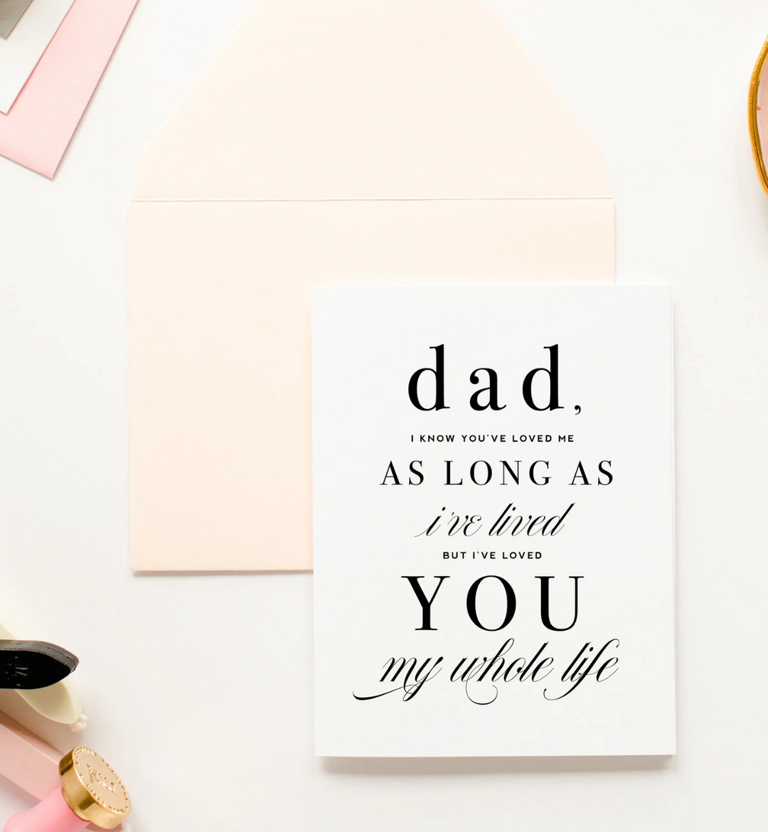 White greeting card with black text that reads "Dad, I know you’ve loved me as long as I’ve lived, but I’ve loved you my whole life." Paired with a light peach envelope and decorative items on a white background