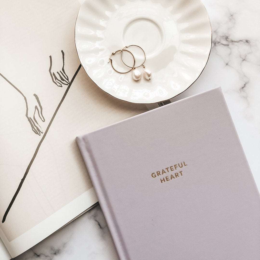 A light purple colored journal entiteld Grateful Heart in gold lettering, a picture of sketched hands above a line, a white saucer with earrings, on a marble table.