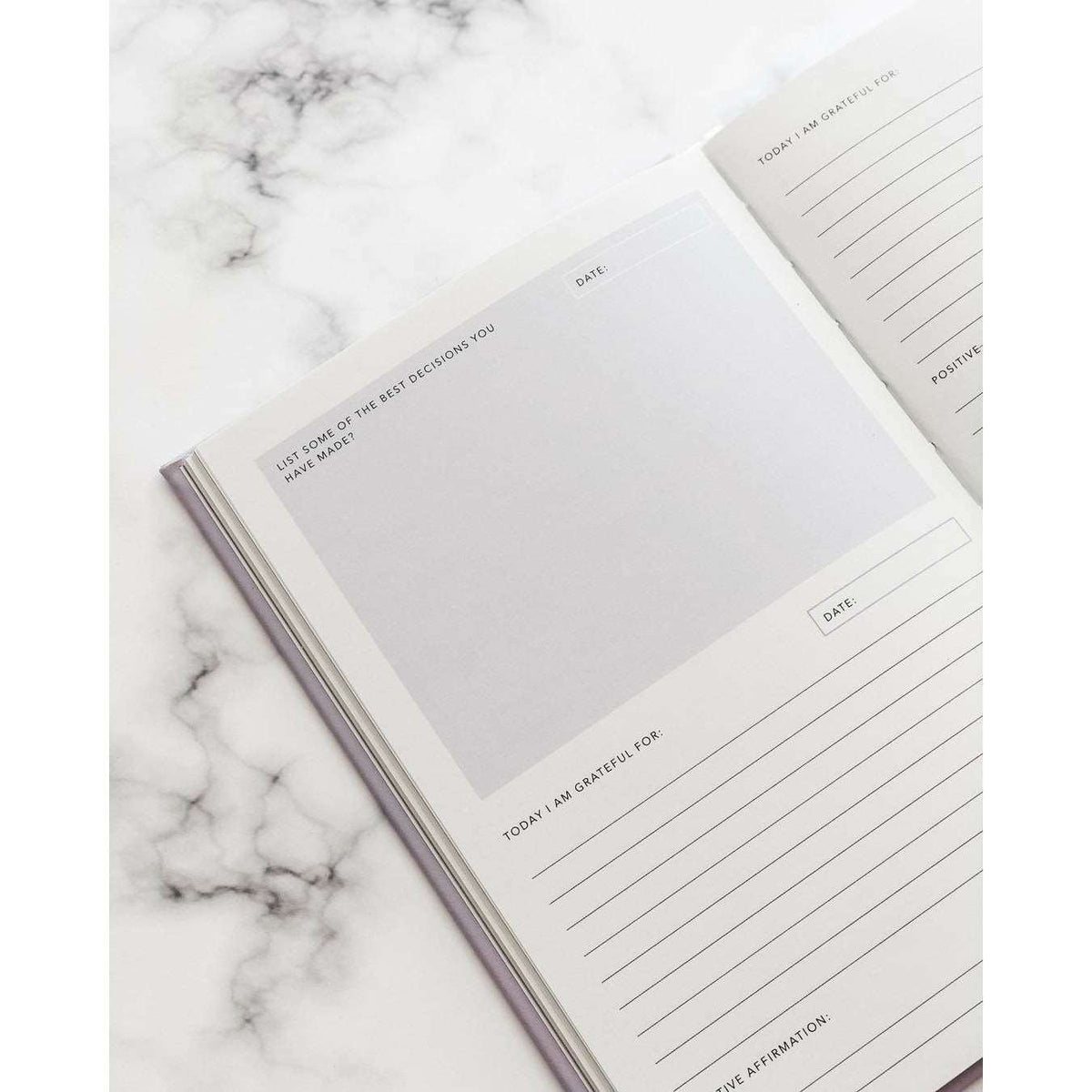 A journal opened to a page with spaces to reply to sections that read: today I am grateful for, the date, and a positive affirmation. The opposite page reads "list of some of the best decisions you have made". On top of a marble table 