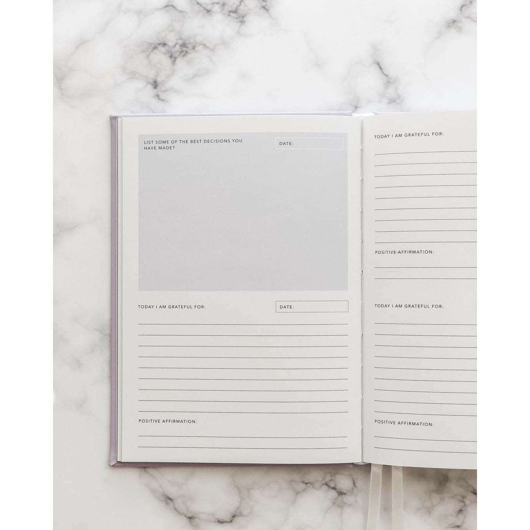 A journal opened to a page with spaces to reply to sections that read: today I am grateful for, the date, and a positive affirmation. The opposite page reads "list of some of the best decisions you have made". On top of a marble table 