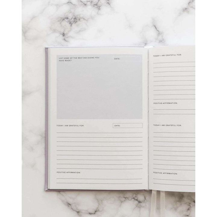 A journal opened to a page with spaces to reply to sections that read: today I am grateful for, the date, and a positive affirmation. The opposite page reads "list of some of the best decisions you have made". On top of a marble table 
