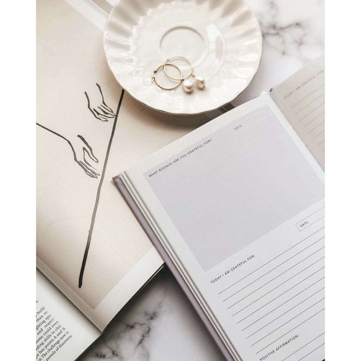 A journal opened to a page that has spaces to reply to sections that read what books are you grateful for, on top of a marble table with a saucer and a cup of coffee. day I am grateful for, and positive affirmations, another book a sketch of hands and a line, white saucer with earrings all on top of a marble table