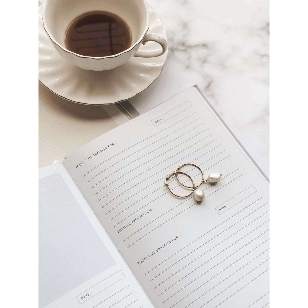 A journal opened to a page with spaces to reply to sections that read: today I am grateful for, the date, and a positive affirmation, on top of a marble table with a saucer and a cup of coffee. 