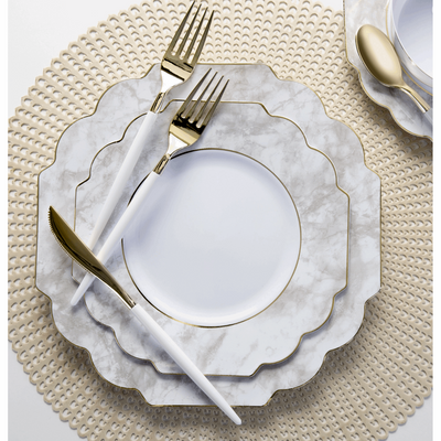 top view of a table setting consisting of a tan placemat with a stacked marble trim white plate set and white handled gold utensils
