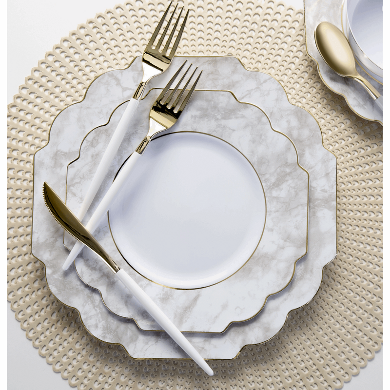 top view of a table setting consisting of a tan placemat with a stacked marble trim white plate set and white handled gold utensils