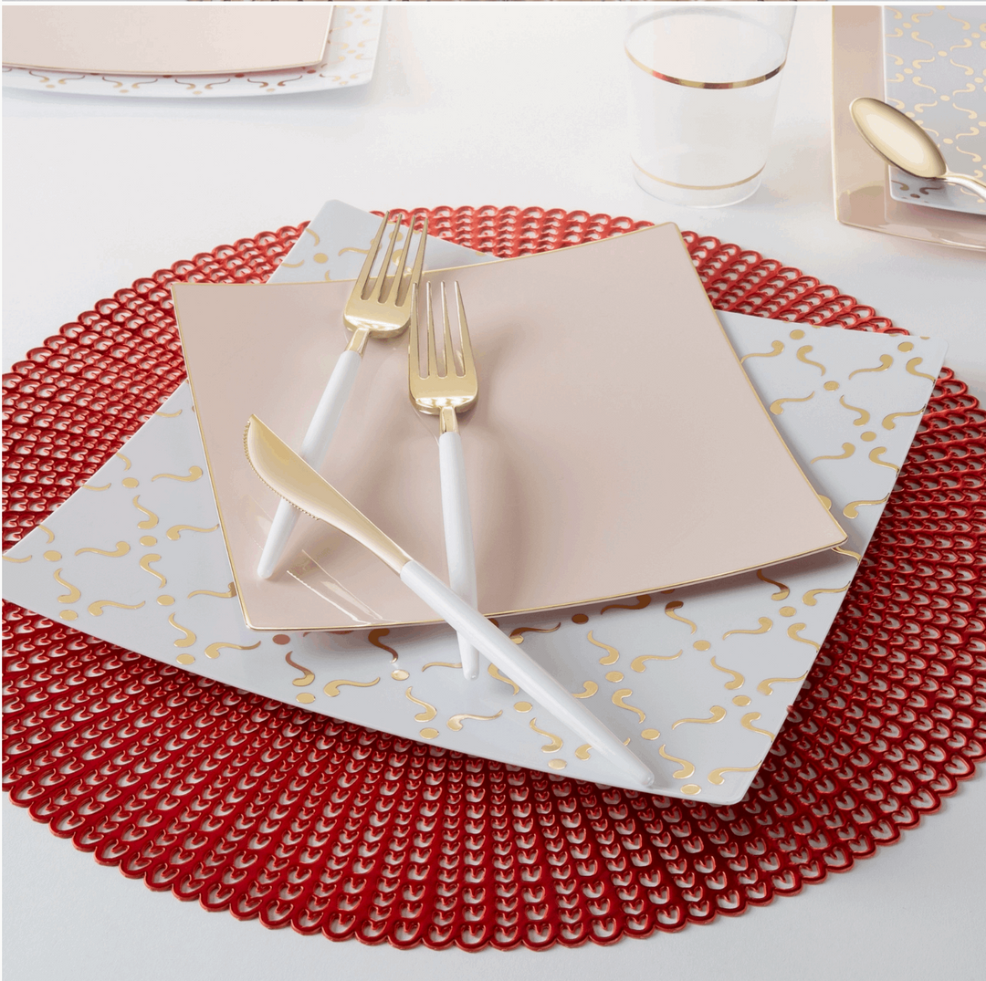 Top-side view of a red placemat, white and gold square dinner plate, blush pink square salad plate, white handled gold utensils, and a clear plastic cup with gold stripe detail.