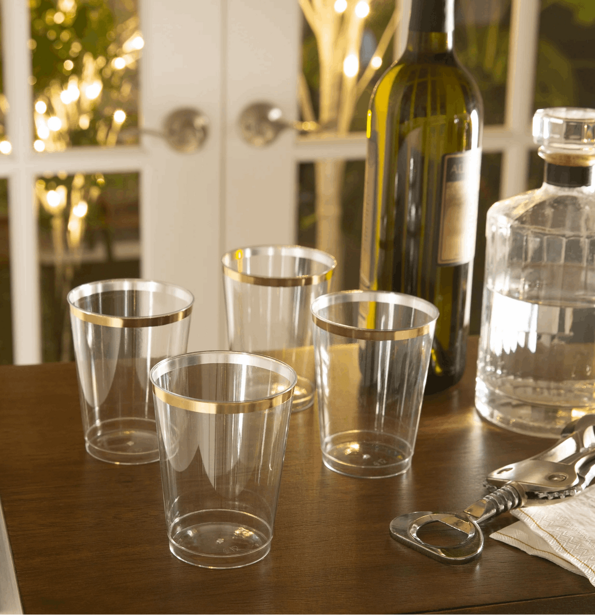 wooden table with 4 clear tumbler style cups with gold rims, a silver bottle opener, bottle of wine, half-filled decanter bottle in front of a window paned white French doors