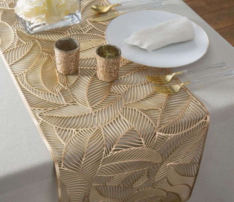 Gold decorative table runner with leaf pattern on a table setting with glitter gold candle holders, a white plate, clear handled gold flatware, and a clear glass vase with cream flowers