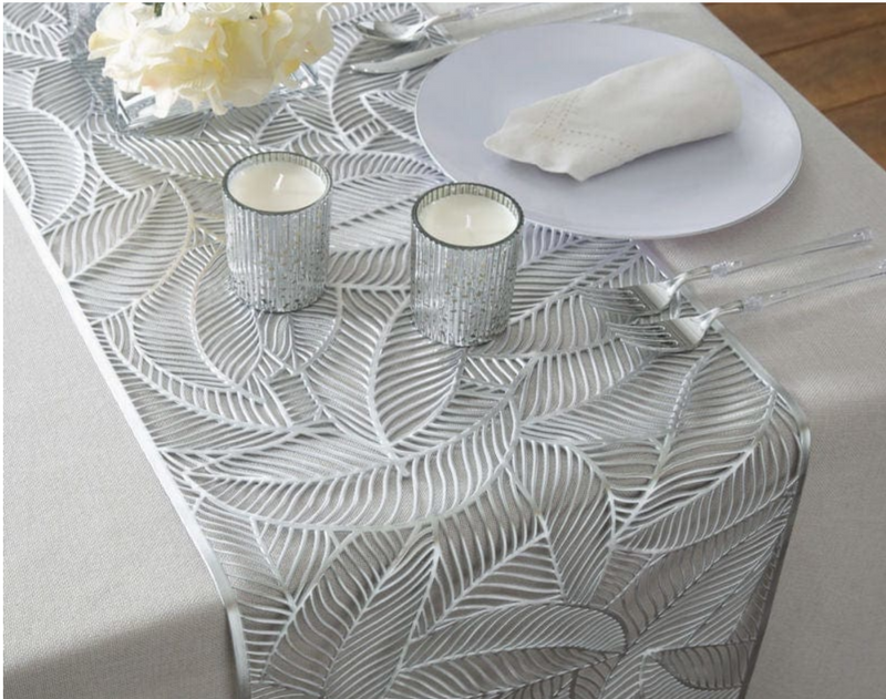 Silver decorative table runner with leaf pattern on a table setting with silver candle holders, a white plate, clear handled silver flatware, and a clear glass vase with cream flowers