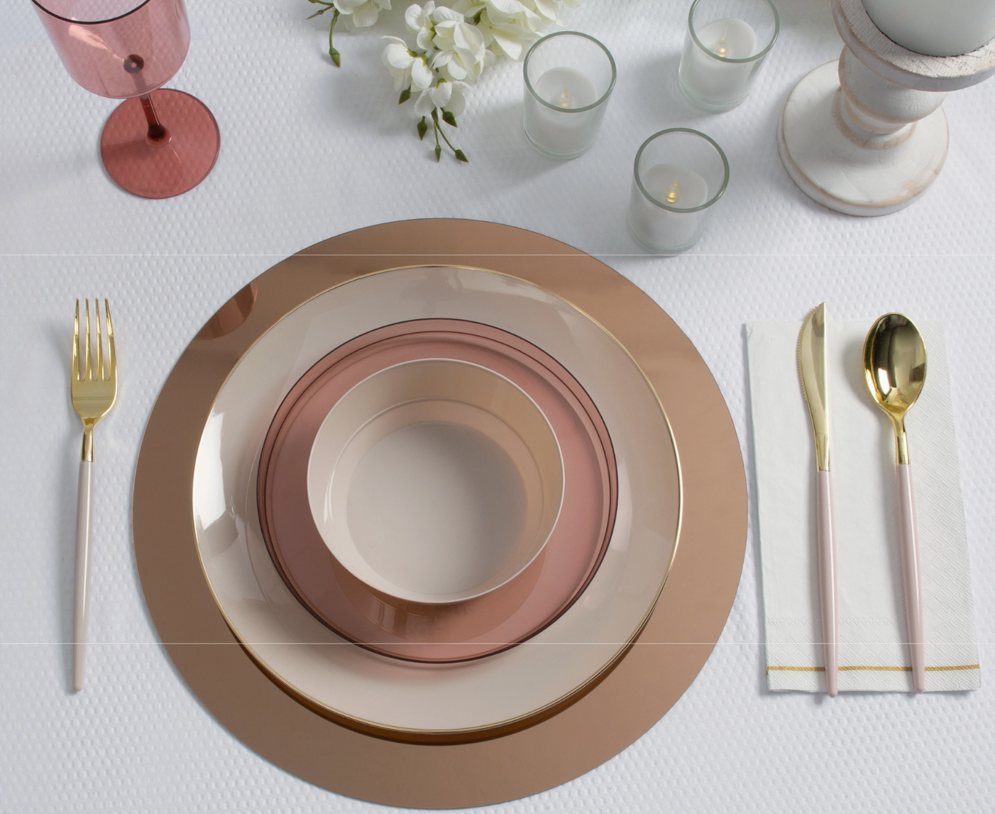 white tablecloth background, mauve colored plastic wine glass, white flowers, 3 votive candles in clear glass votive vases. rose gold charger plate, topped with a gold rimmed cream dinner plate, mauve colored dessert plate and a cream salad bowl, flanked on either side with linen color handle gold utensils.
