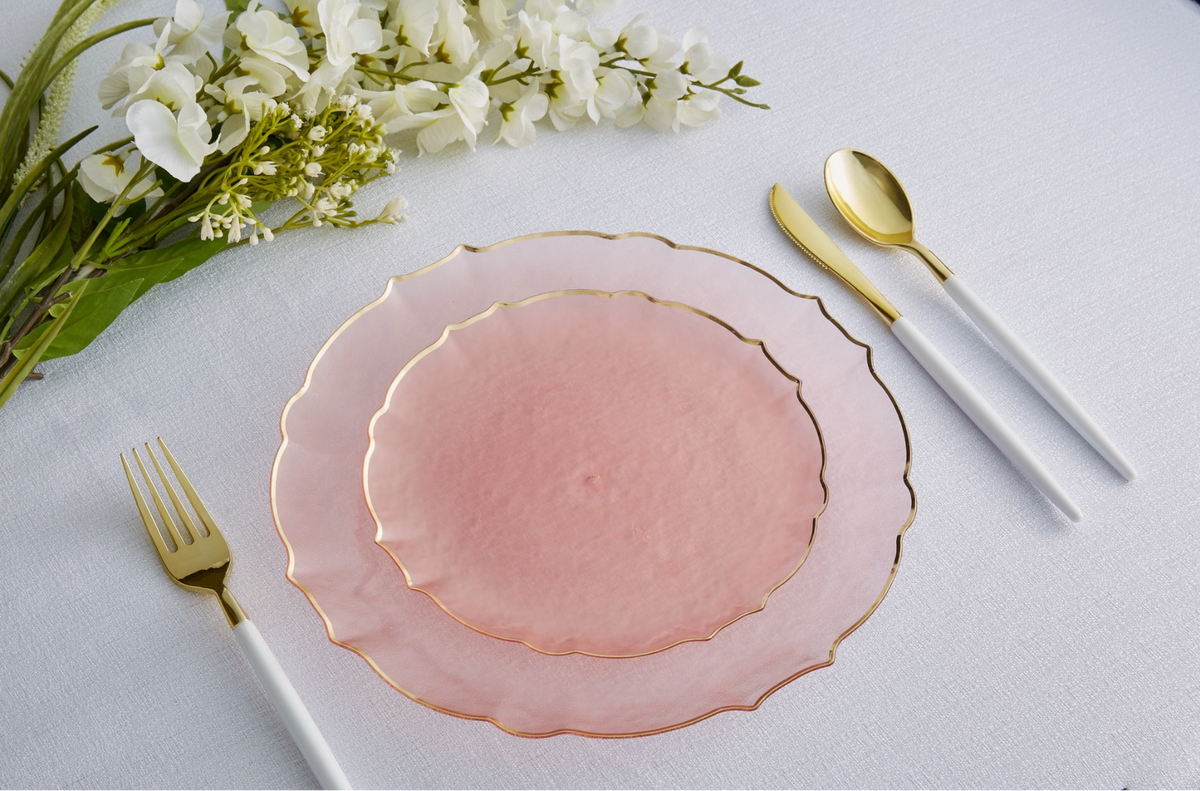 Pink frosted finish plastic dinner and salad plates with gold rim on a white tablecloth, accompanied by white and gold fork, spoon, and knife set and white flowers.