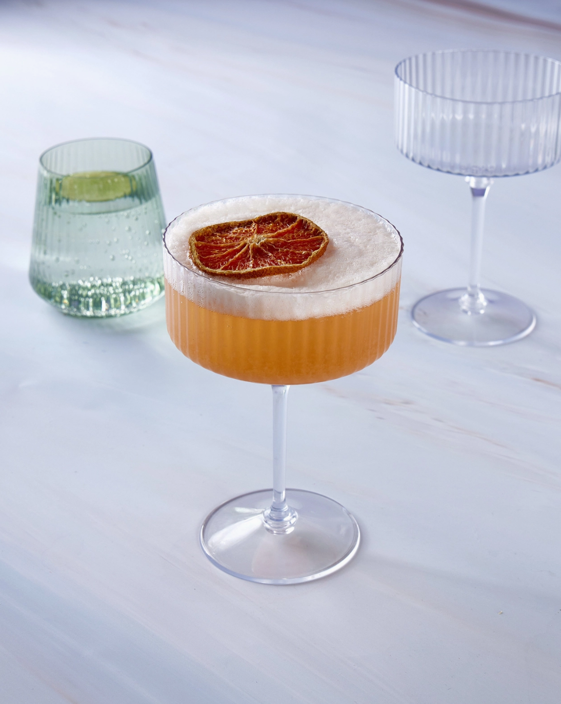 2 plastic clear ribbed coupe shaped champagne glass, 1 filled with an orange beverage and a dried orange slice in it, and 1 mint green colored plastic stemless goblet style glass filled with sparkling water and a lime slice in it on a white marble background tabletop