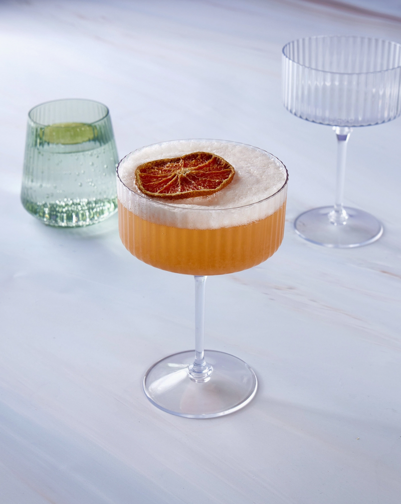 2 plastic clear ribbed coupe shaped champagne glass, 1 filled with an orange beverage and a dried orange slice in it, and 1 mint green colored plastic stemless goblet style glass filled with sparkling water and a lime slice in it on a white marble background tabletop