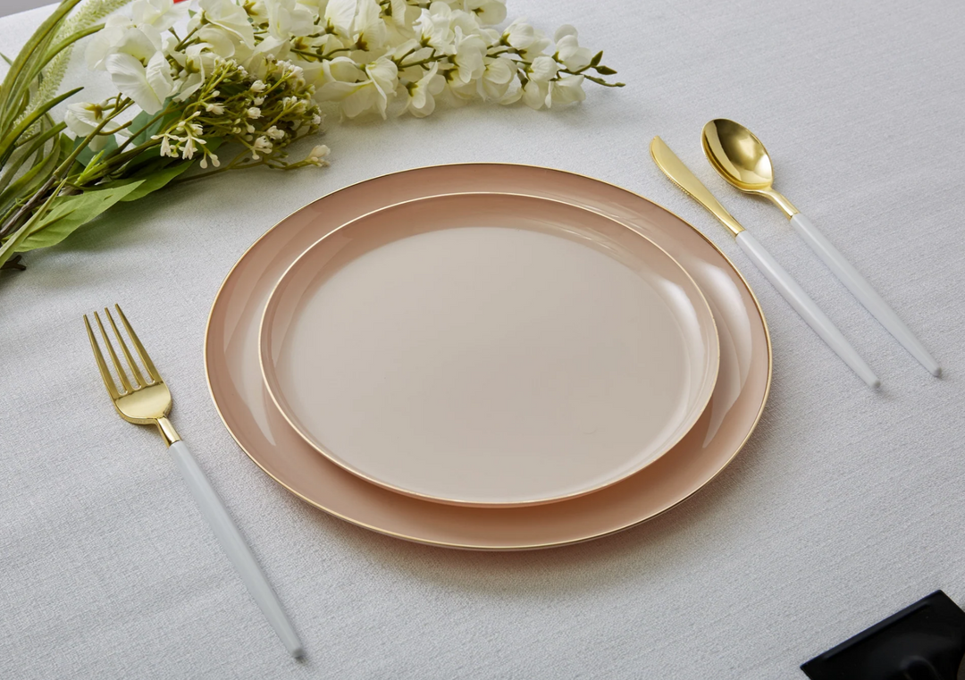 Table setting with pinkish beige dinner and salad plates, white and gold cutlery, and white flowers on a light gray tablecloth surface.