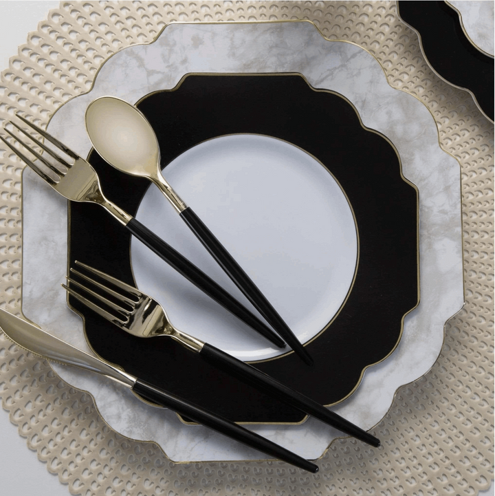 white background with a pale gold placemat topped with a white marble with gold rim charger plate topped with a white with black trim and gold rim dinner plate topped with black handled and gold knife, forks, and spoon.