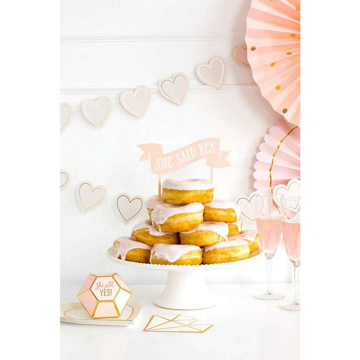 Bridal Shower setting with 2 pink and gold heart shaped banners and 2 pink paper fans on a white wall, a cake stand with pink frosted donuts on it and a small pink banner that says "she said yes", 2 champagne glasses filled with pink wine and diamond shaped favor box that says she said yes in gold foil.