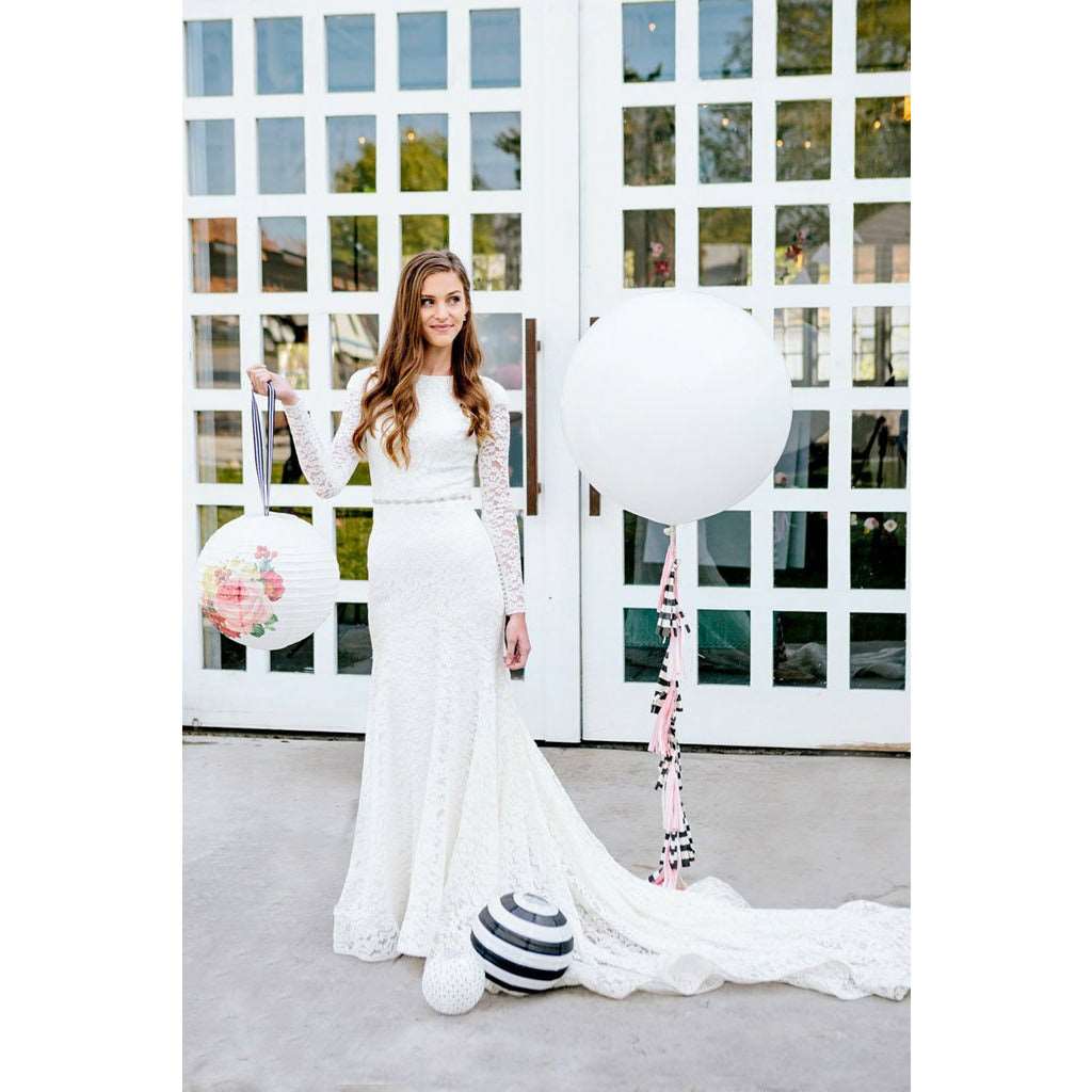 Woman in a wedding dress holding a decorative paper lantern with floral print design and black and white striped ribbon hanger, standing next to a large floating white balloon with pink, black and white ribbon, and a black and white striped balloon all in front of 2 large glass doors