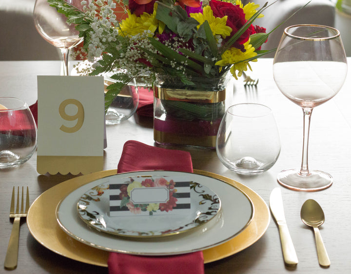 Dining table setting with a floral centerpiece, plates, and cutlery on a wooden table. black and white striped and floral print place card on top of a dinner plate, and a white and gold table number card.