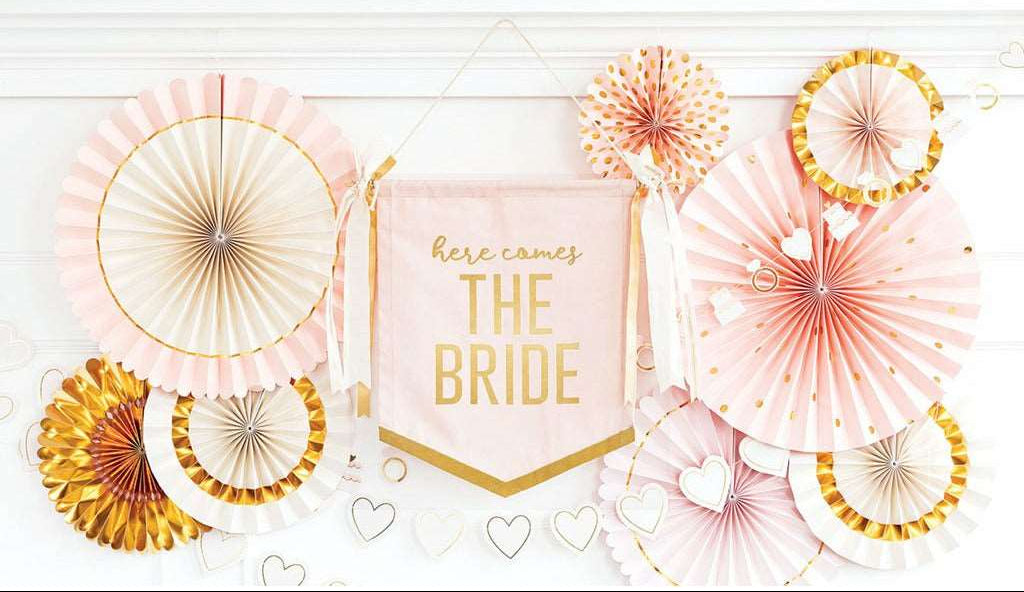 A canvas wall banner with the text 'here comes the bride' in gold lettering, surrounded by decorative fans in gold and pink shades, and a die-cut heart banner intended for bridal shower decorations.