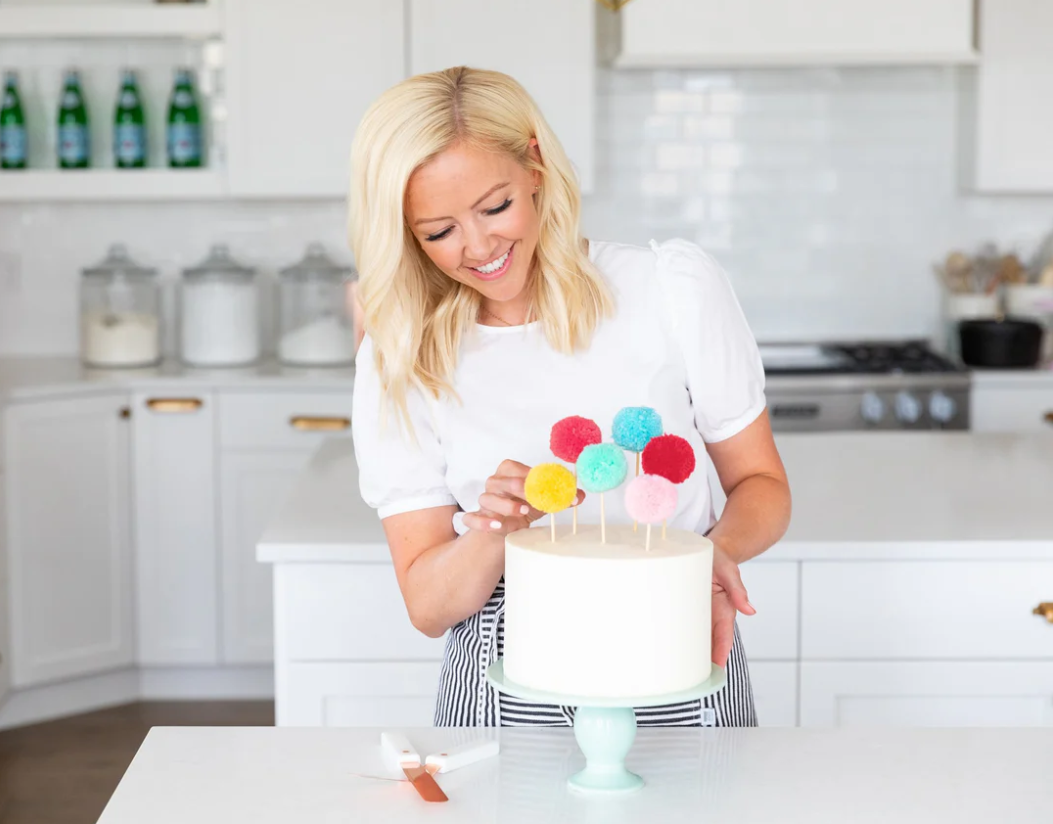 Woman in a kitchen decorating a cake with colorful pom-pom style cake toppers